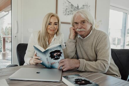 Petra Mattfeldt und Axel Petermann sitzen an einem Tisch und zeigen ihr gemeinsames Buch in die Kamera. Auf dem Tisch außerdem ein Laptop und ein weiteres Buch.