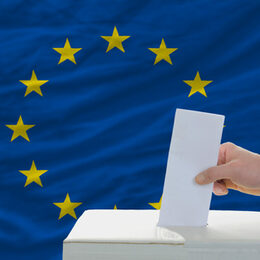 Eine Hand steckt ein Stück Papier in eine Wahlurne. Im Hintergrund die Europäische Flagge.